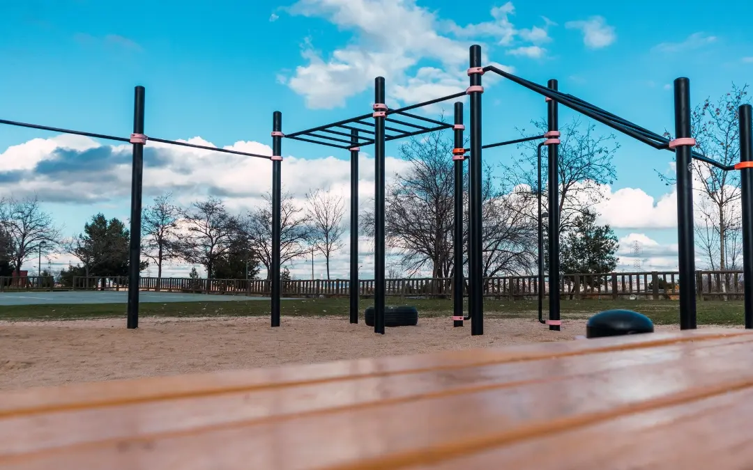 Aire de street workout en extérieur avec barres de traction, échelle horizontale et pneus posés sur le sable, vue depuis un banc en bois